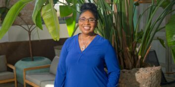 a photo of sharon chambers gordon in a blue shirt smiling in front of some plants and furniture