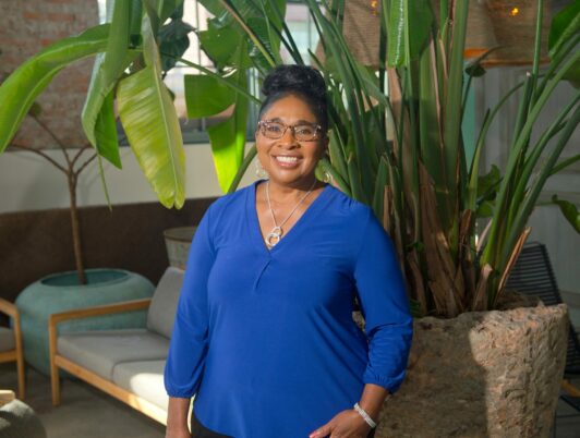 a photo of sharon chambers gordon in a blue shirt smiling in front of some plants and furniture
