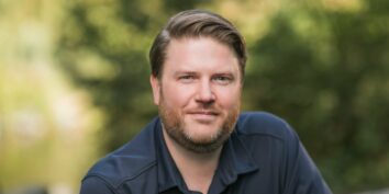 erik hanberg poses in a blue shirt in front of some greenery in tacoma