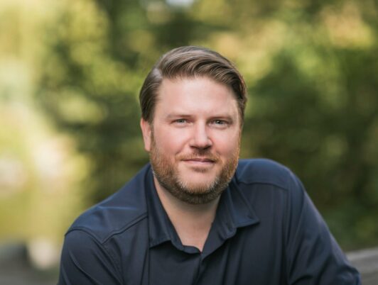 erik hanberg poses in a blue shirt in front of some greenery in tacoma
