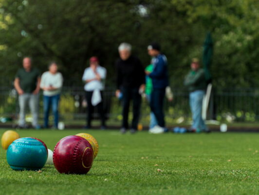 Three lawn bowling balls in the foreground with people from the Tacoma Lawn Bowling Club bowling across a green lawn in the distance.