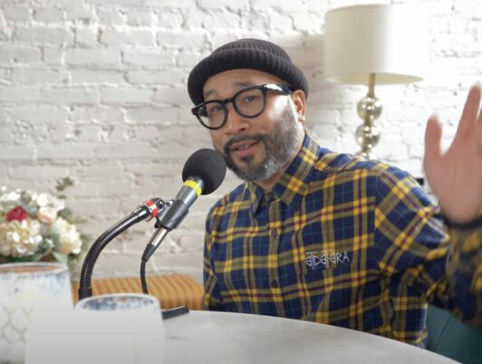 Silong Chhun wears a beanie and glasses with a plaid shirt while gesturing and speaking into a podcast mic on the Move to Tacoma Podcast.