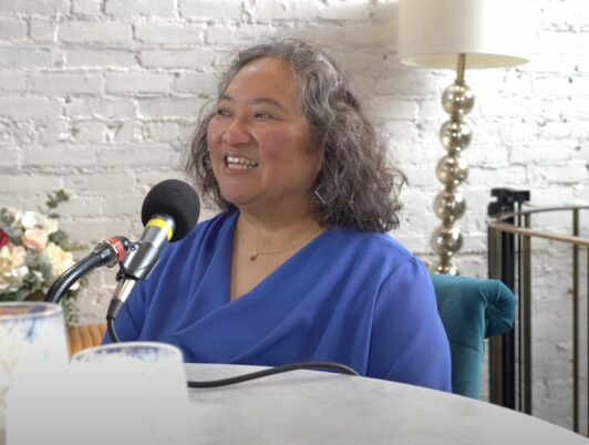 Author Tamiko Nimura sits at a table with a podcast mic in a blue shirt.